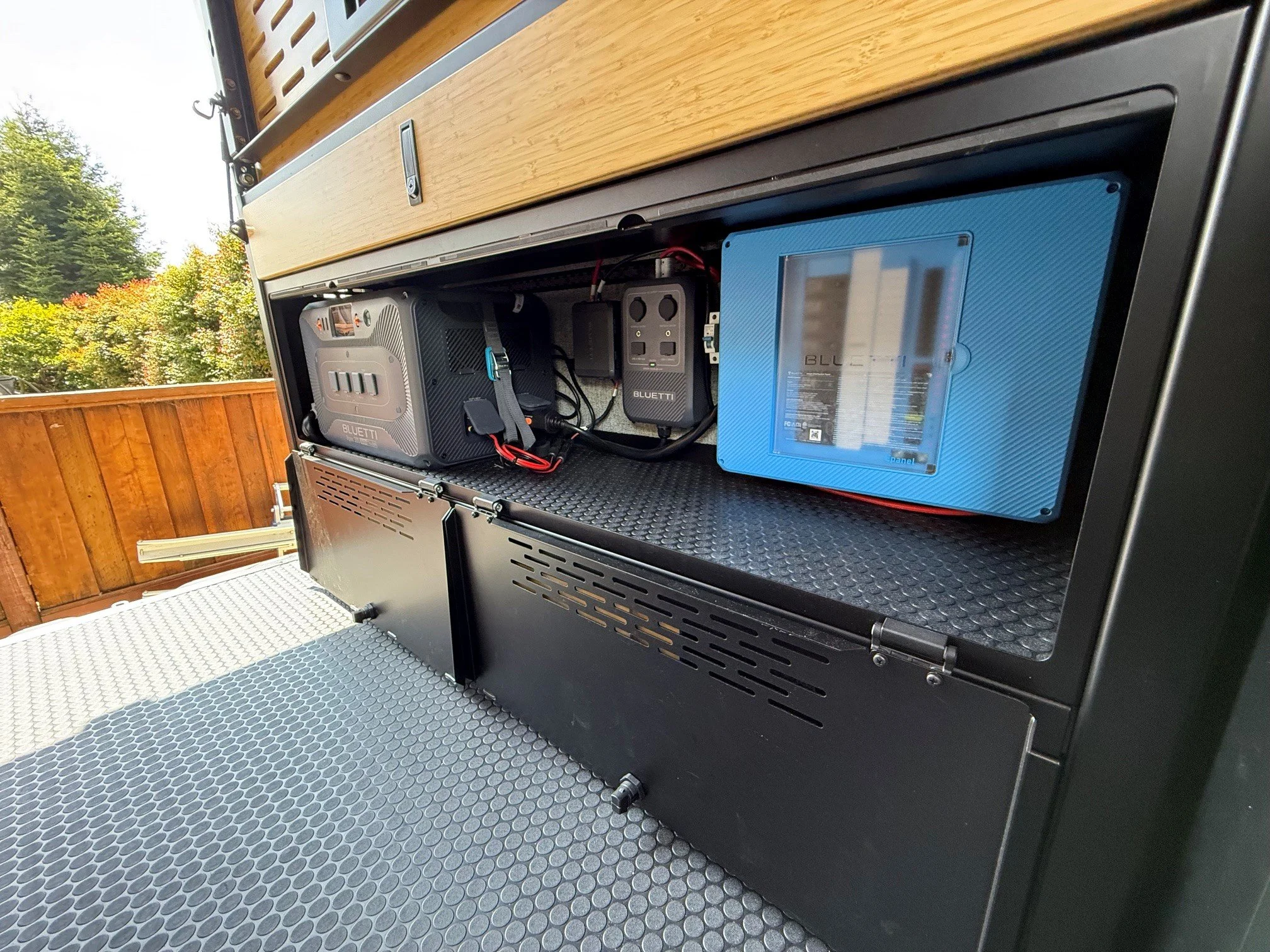 Image shows the interior power supply, and storage compartments with a textured rubber flooring, set against a backdrop of trees and a wooden fence.