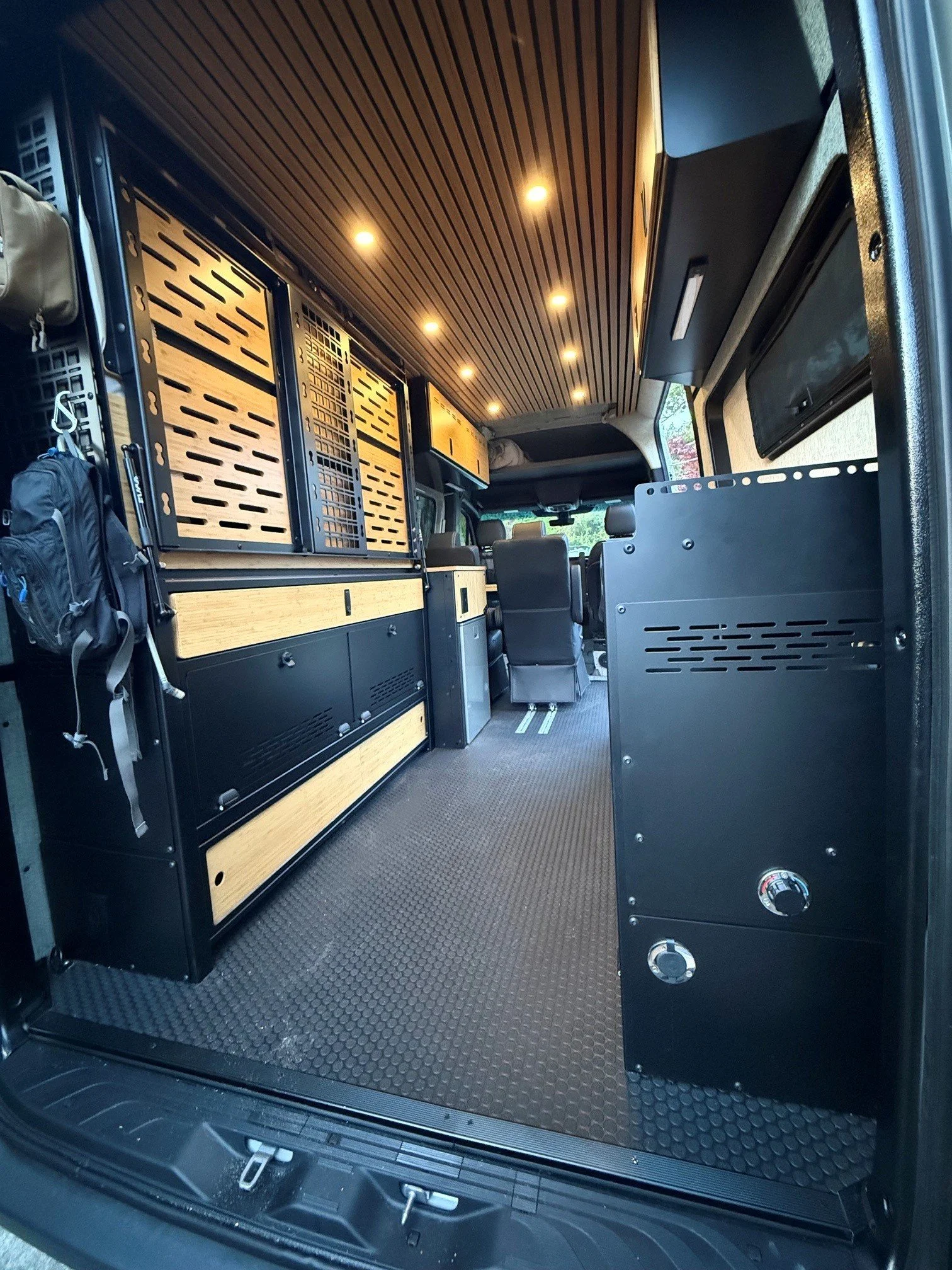 Interior of a converted van featuring wooden storage compartments, a textured floor, and a small black stove with control knobs.