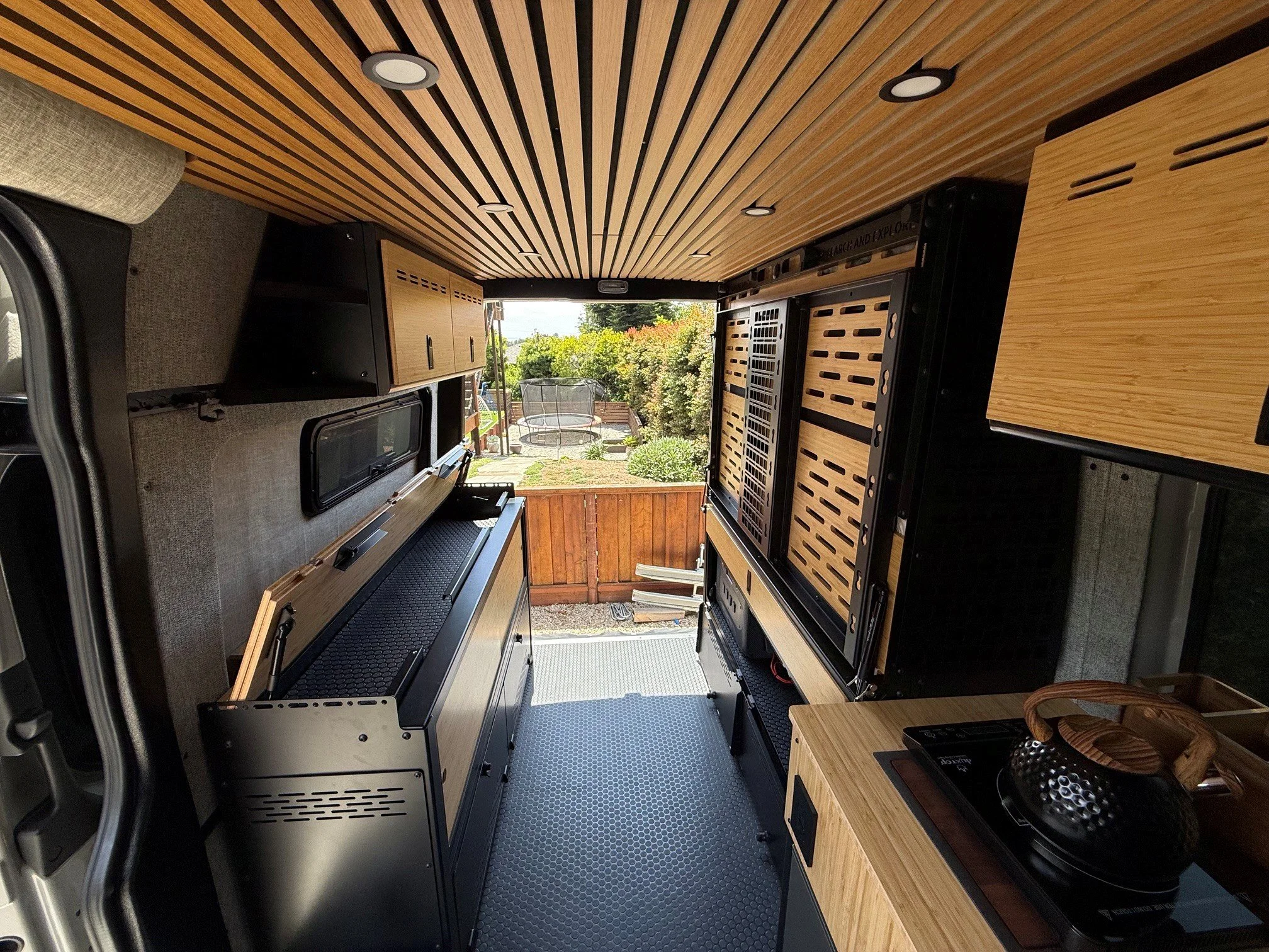 Interior of a converted van with wood-paneled ceiling and walls, showing a small kitchen area with a stove, countertop, and storage cabinets, overlooking a backyard with trees and a deck.