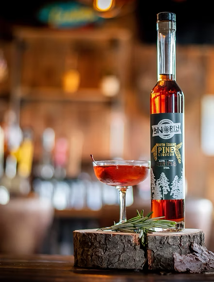 A bottle of North Idaho Pine Liqueur and a cocktail glass filled with red liquid on a wooden surface with a sprig of rosemary, in a bar setting.