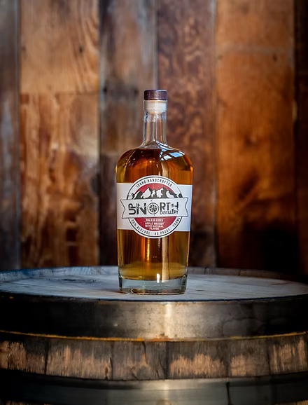 A glass bottle of whiskey with a label, placed on a wooden barrel in a rustic setting.