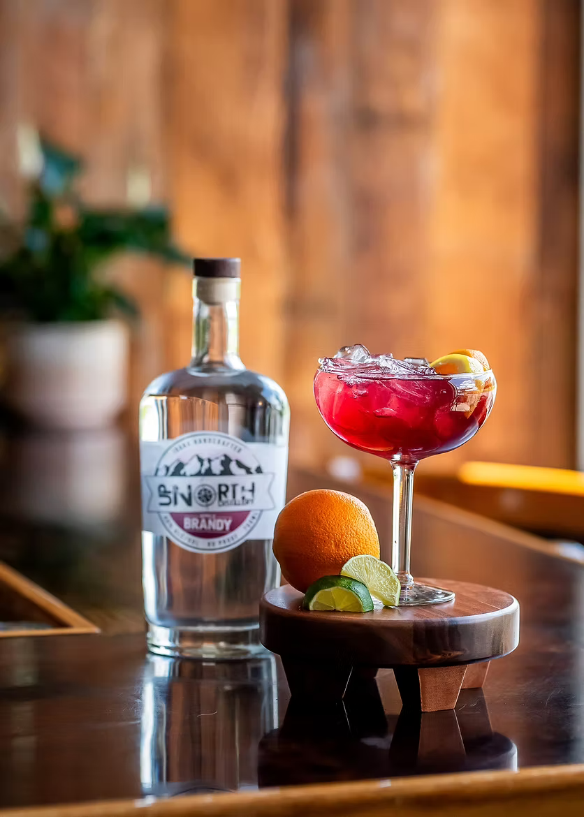 A bottle of brandy, a glass cocktail with red liquid and crushed ice garnished with lemon slices, an orange, and lime slices on a wooden table with a blurred wooden wall background.