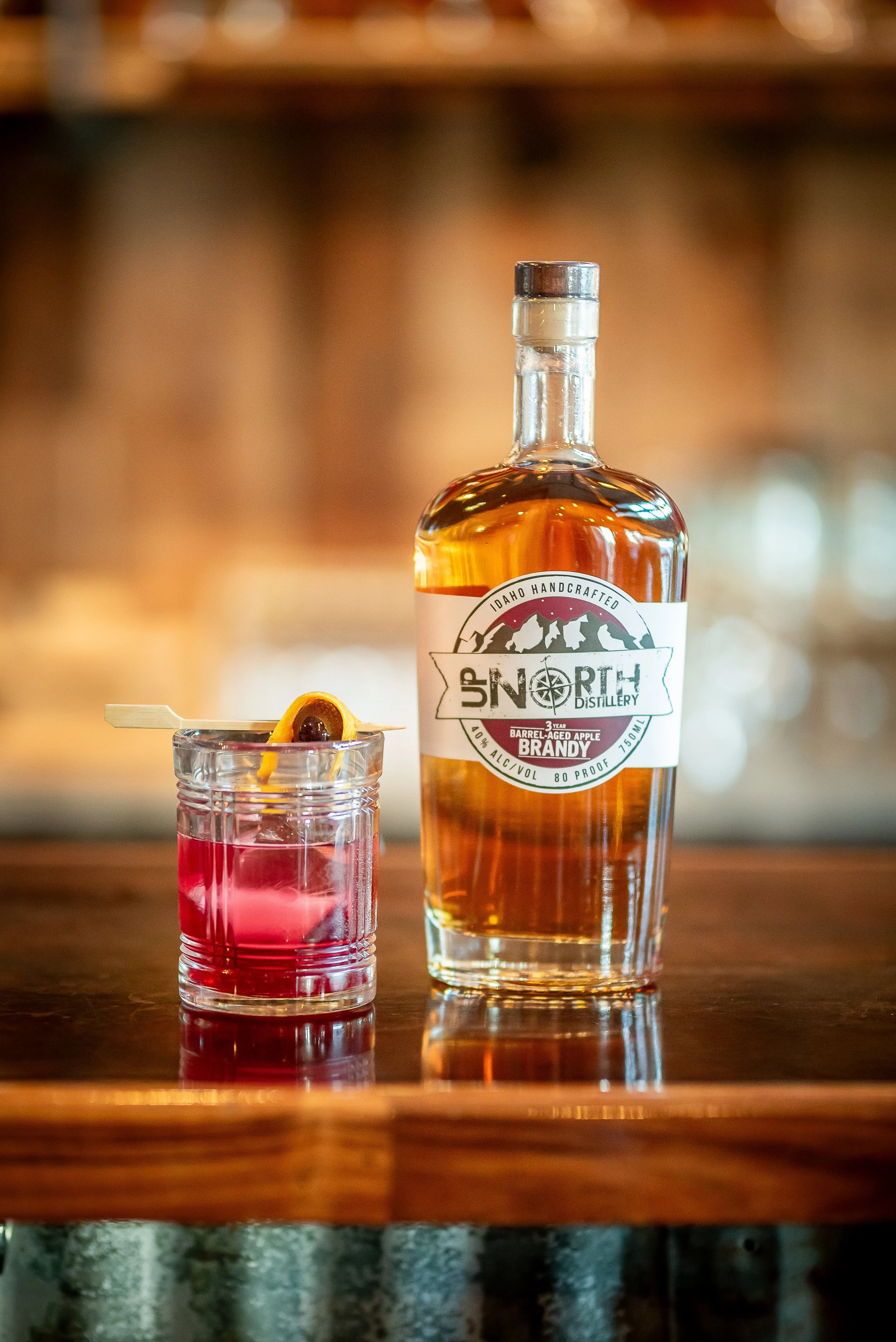A bottle of Idaho handcrafted apple brandy and a pink cocktail garnished with a cherry and a lemon twist on a wooden bar
