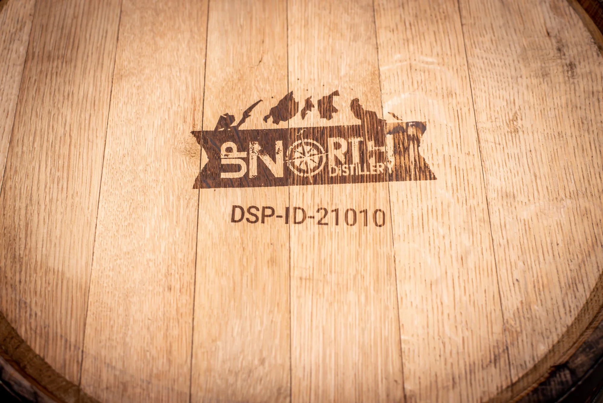 Wooden barrel with a logo that reads 'SNOWHITE DISTILLERY' and includes a mountain silhouette and a compass, along with a code 'DSP-ID-21010'.