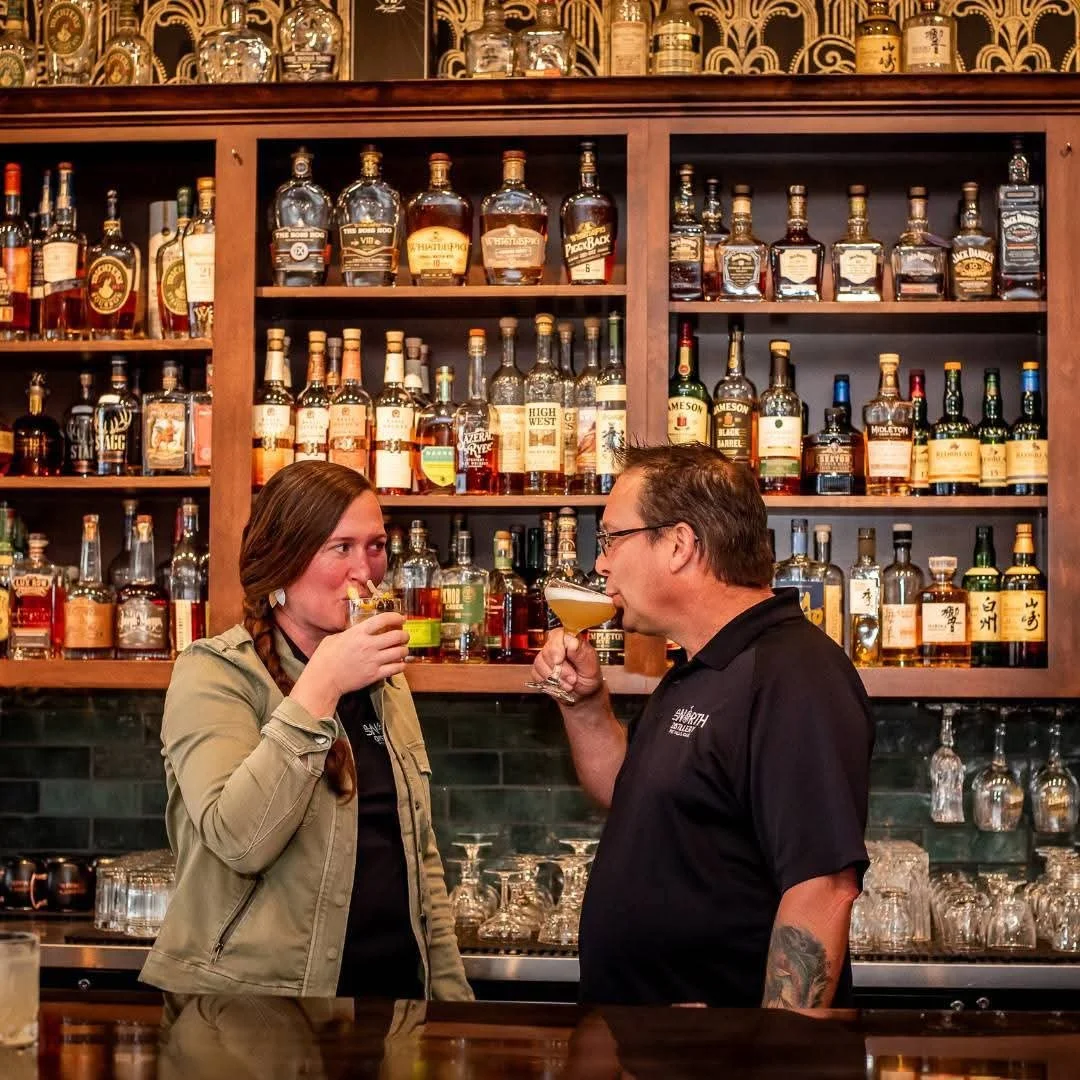 A man and woman clinking glasses and drinking cocktails in a bar with a wooden shelf filled with liquor bottles behind them.