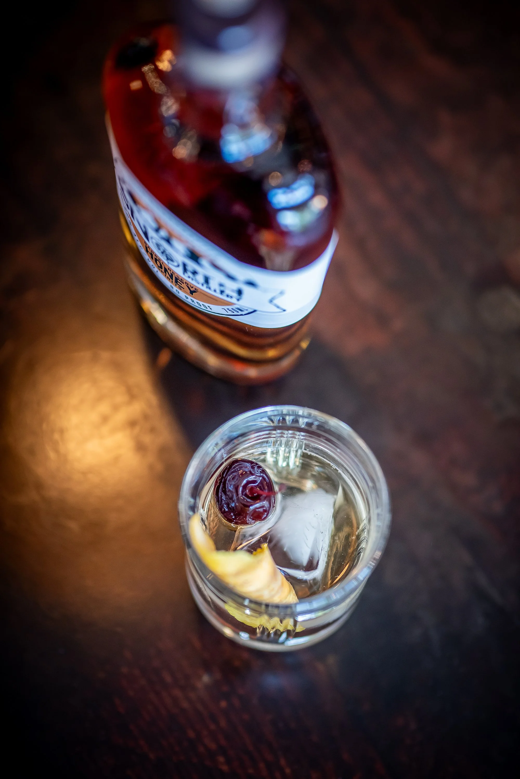 A bottle of Hennessy Cognac placed on a dark wooden surface and a glass of ice with a lemon slice and a cherry.