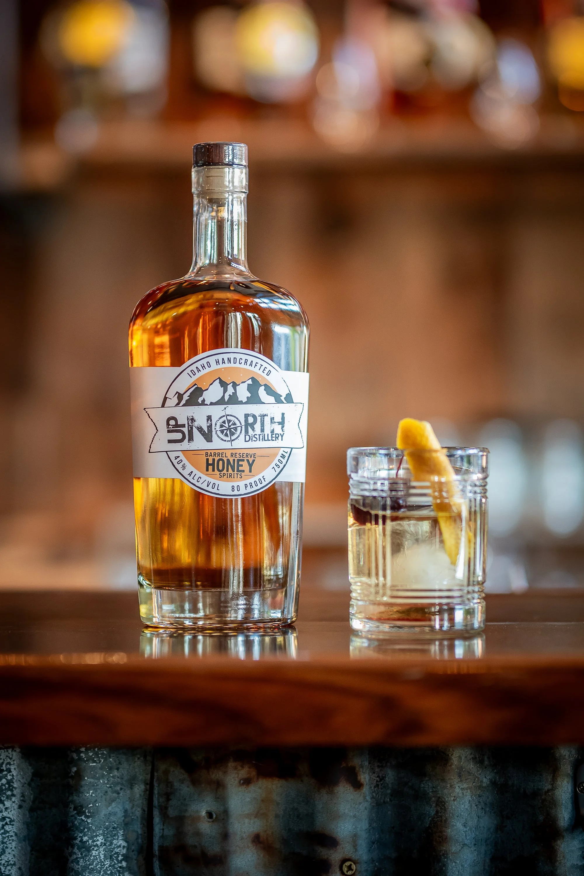 A bottle of honey spirit with a mountain logo and a glass with ice and a lemon twist on a wooden bar counter.