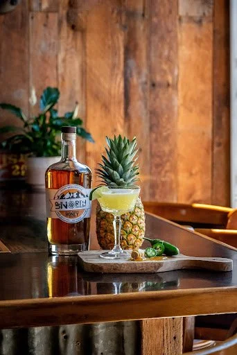 A bottle of honey liqueur, a pineapple, and a margarita glass with lime and pineapple garnishes on a wooden table with a wooden wall in the background.