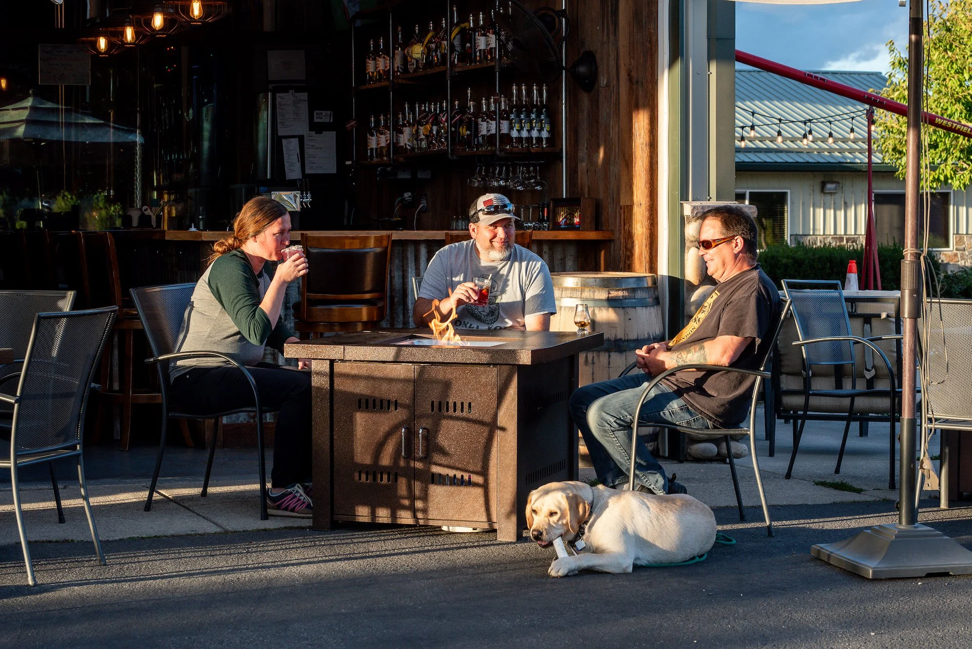 Three people sitting around a fire pit outside a restaurant, drinking beverages, with a yellow Labrador dog lying nearby. The background shows a wooden bar area inside the restaurant and a building with a corrugated metal roof outside, with trees and string lights visible.