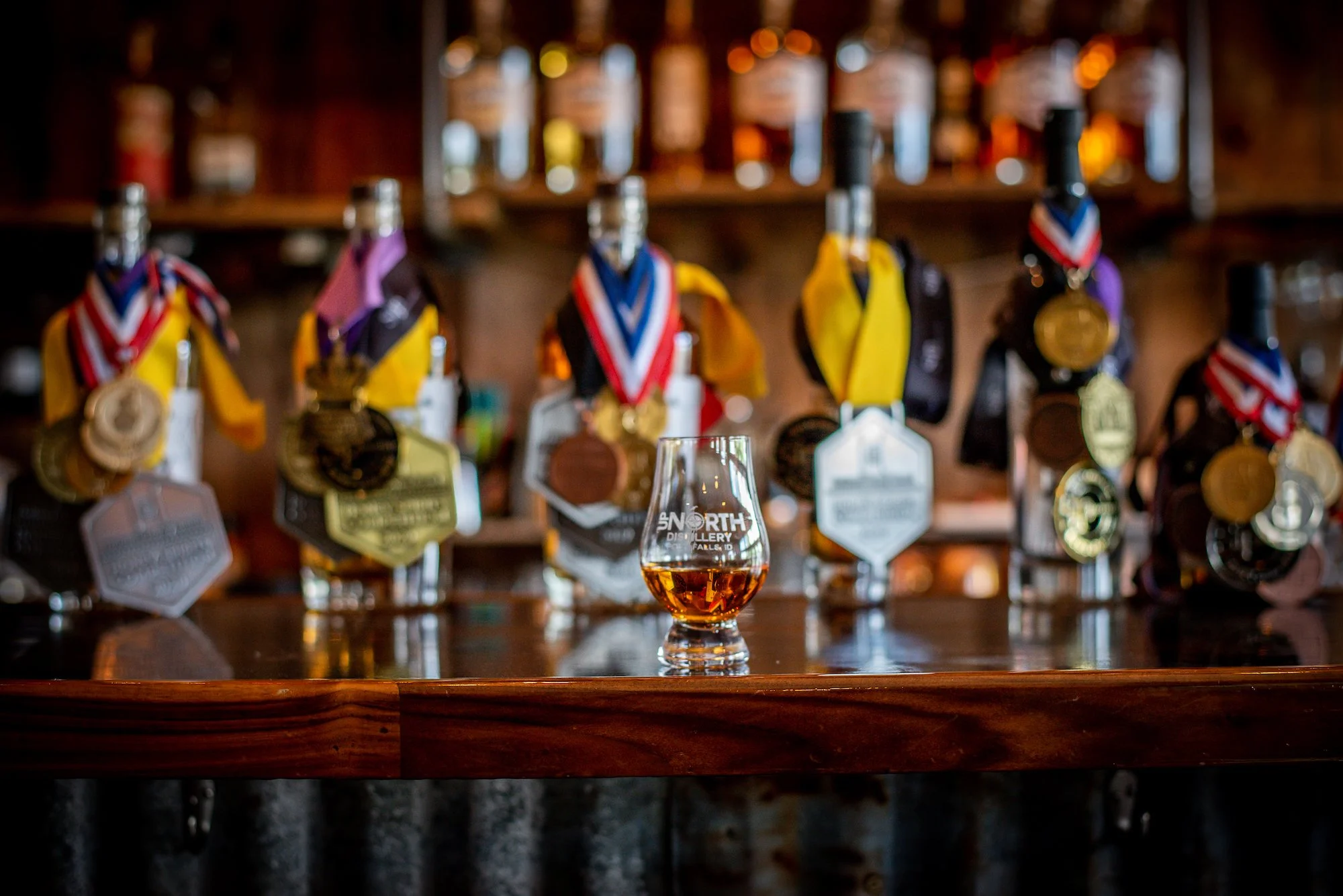 Bar with a glass of whiskey in the foreground and multiple bottles of whiskey on shelves in the background, some with medals and awards.