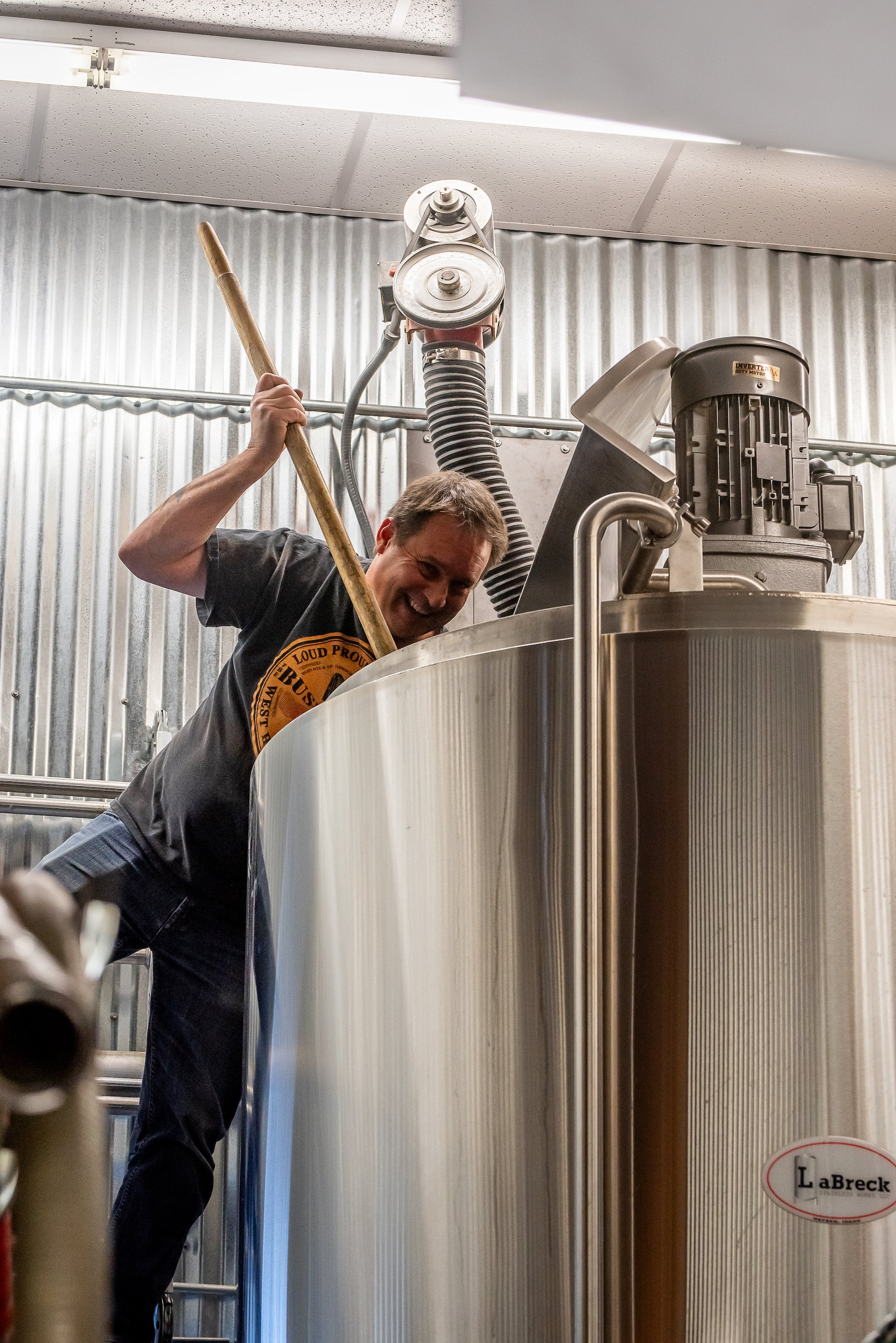 A person smiling and holding a long stick in front of a large stainless steel brewing tank inside an industrial or brewery setting.
