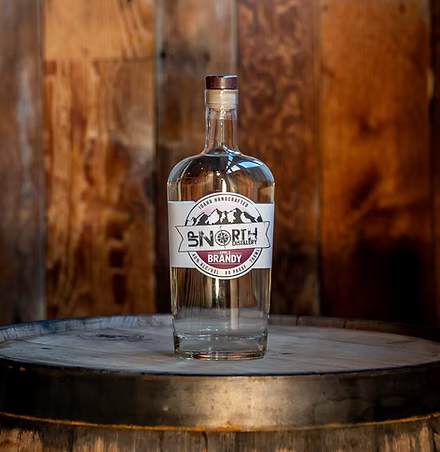 A clear glass bottle of Brandy with a label reading "SOUTH DISABILITY BRANDY" placed on a wooden barrel with a metal band, set against a rustic wooden background.