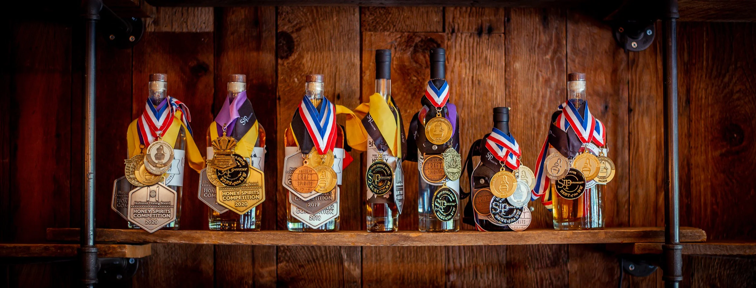 Bottles of honey spirits on a wooden shelf, each with medals hanging around necks, awarded in honey spirits competitions from 2018 to 2020.