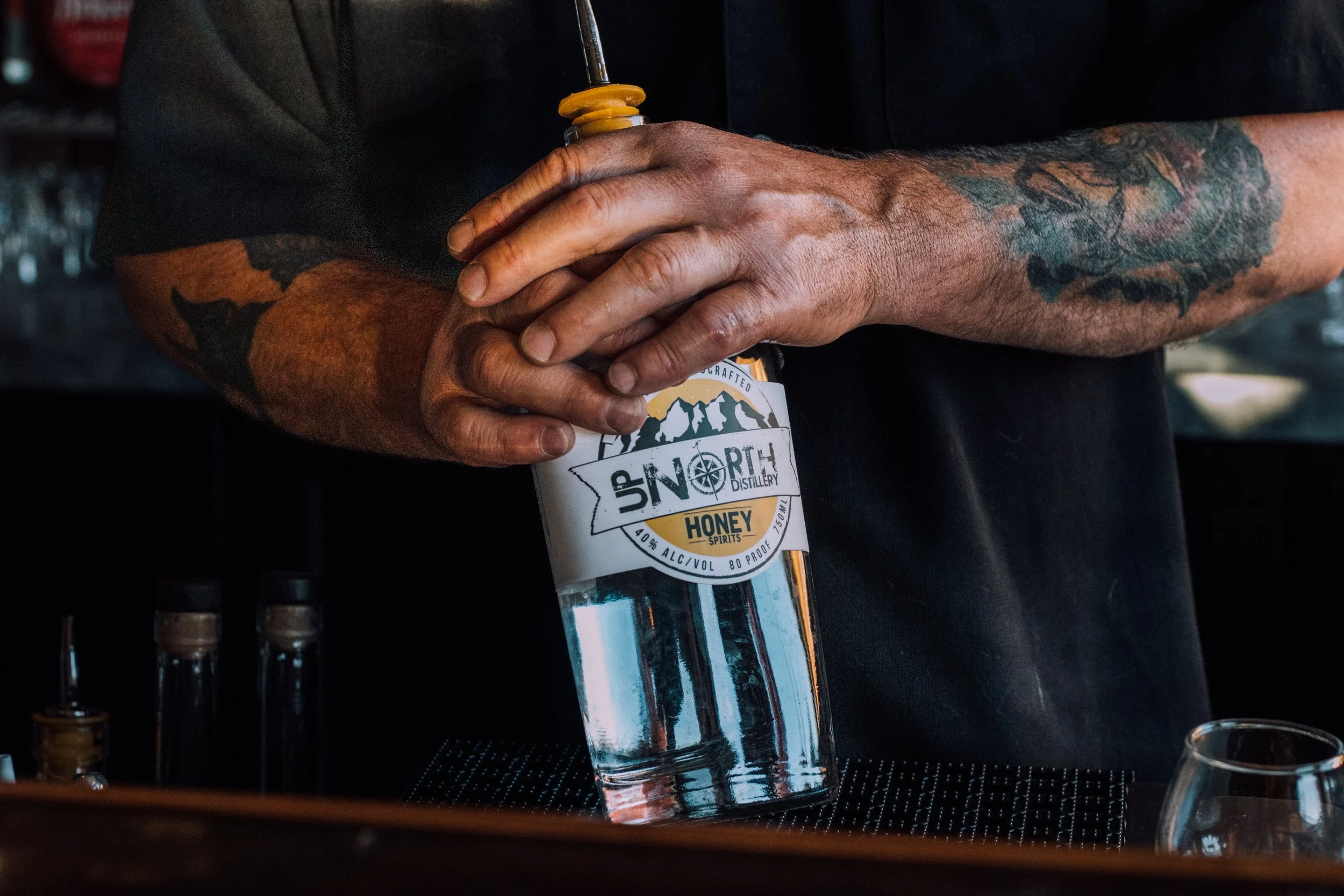 A hand holding a bottle of UP NORTH Honey Spirit, with visible tattoos on the arm, in what appears to be a bar or distillery setting.