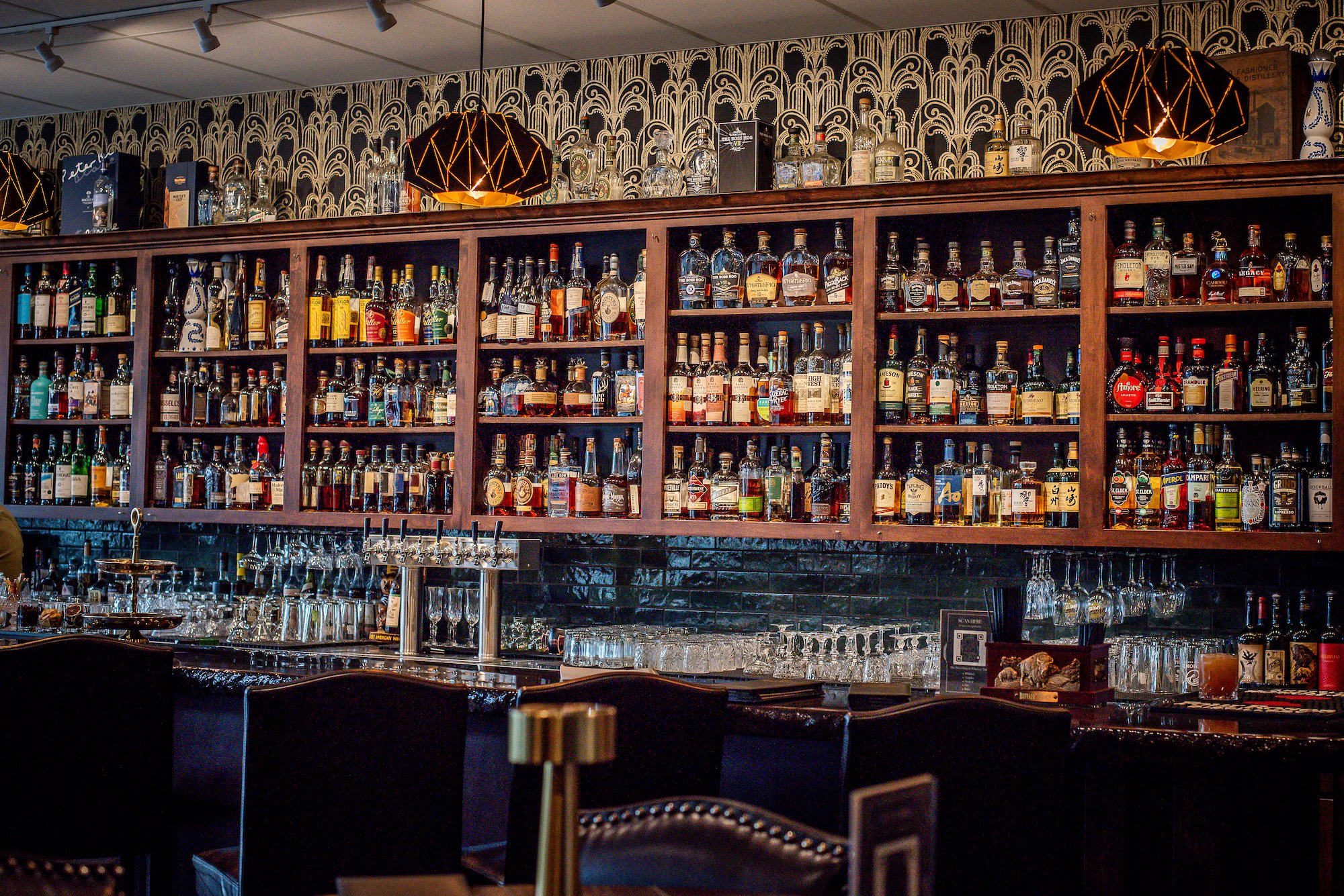 A bar with a wooden shelf holding various bottles of liquor and alcohol, illuminated by warm lighting with decorative black and gold lamps hanging above, and a patterned wallpaper background.