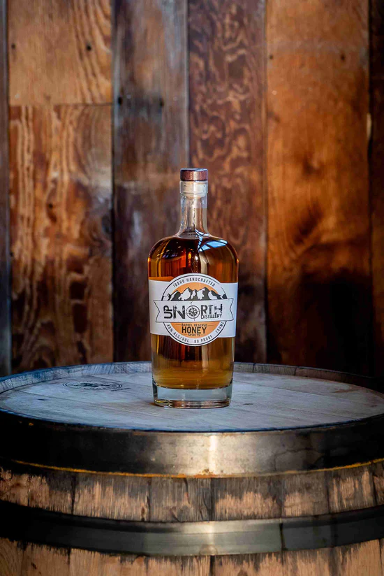 A bottle of North Honey bourbon whiskey on a wooden barrel against a rustic wood wall background.