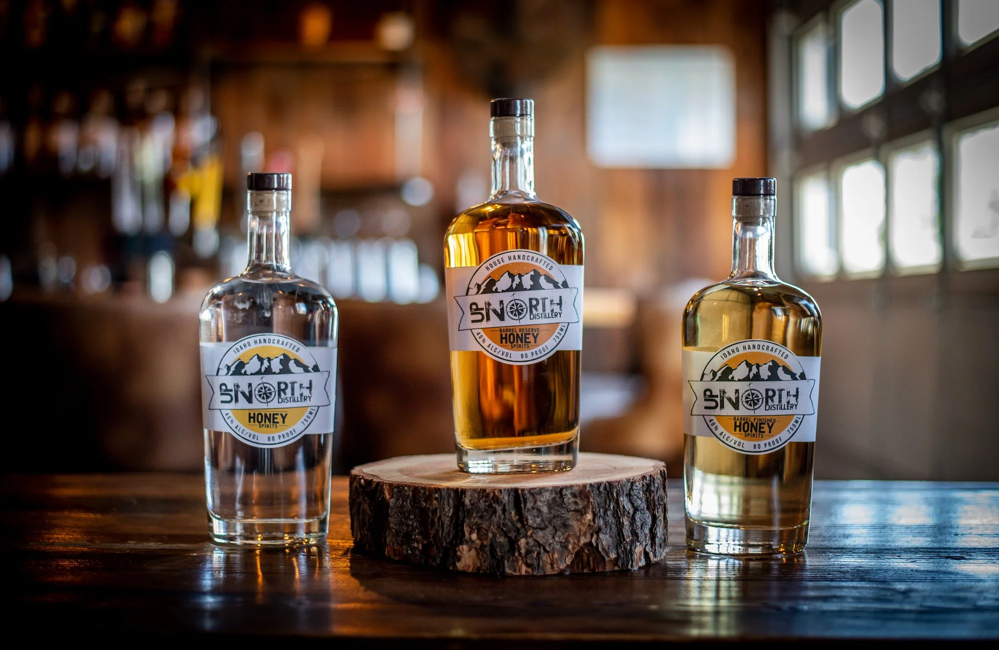 Three bottles of Idaho handcrafted honey spirits on a wooden table, with one bottle in the center placed on a tree stump, inside a rustic bar or brewery with wooden walls and large windows.
