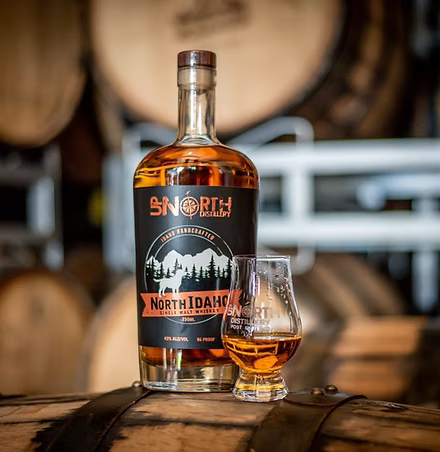 A bottle of North Idaho whiskey and a glass of whiskey on a wooden barrel in a distillery setting.