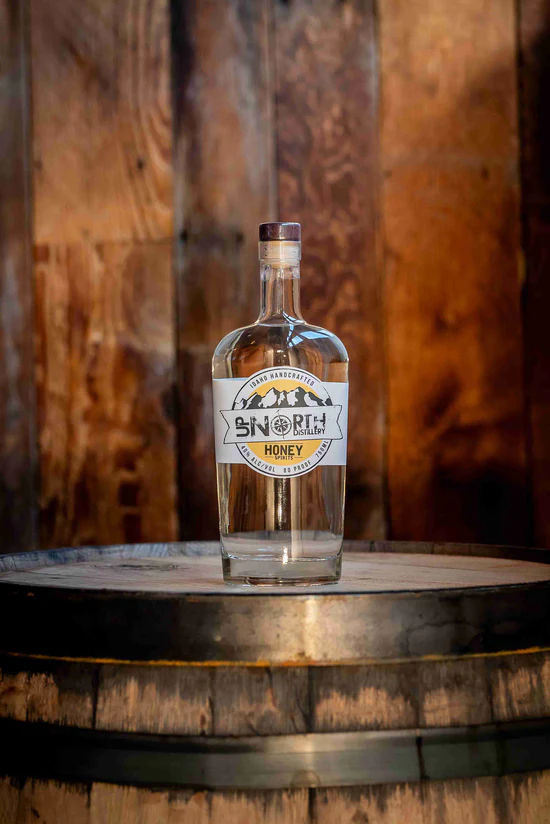 A glass bottle of honey-flavored vodka from North Craft Distillery, placed on a wooden barrel in a rustic setting with wooden walls.