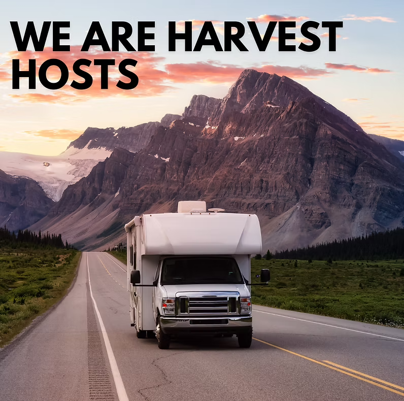 A white RV driving on an empty road with mountains in the background during sunset, with text overlay saying 'We Are Harvest Hosts'.