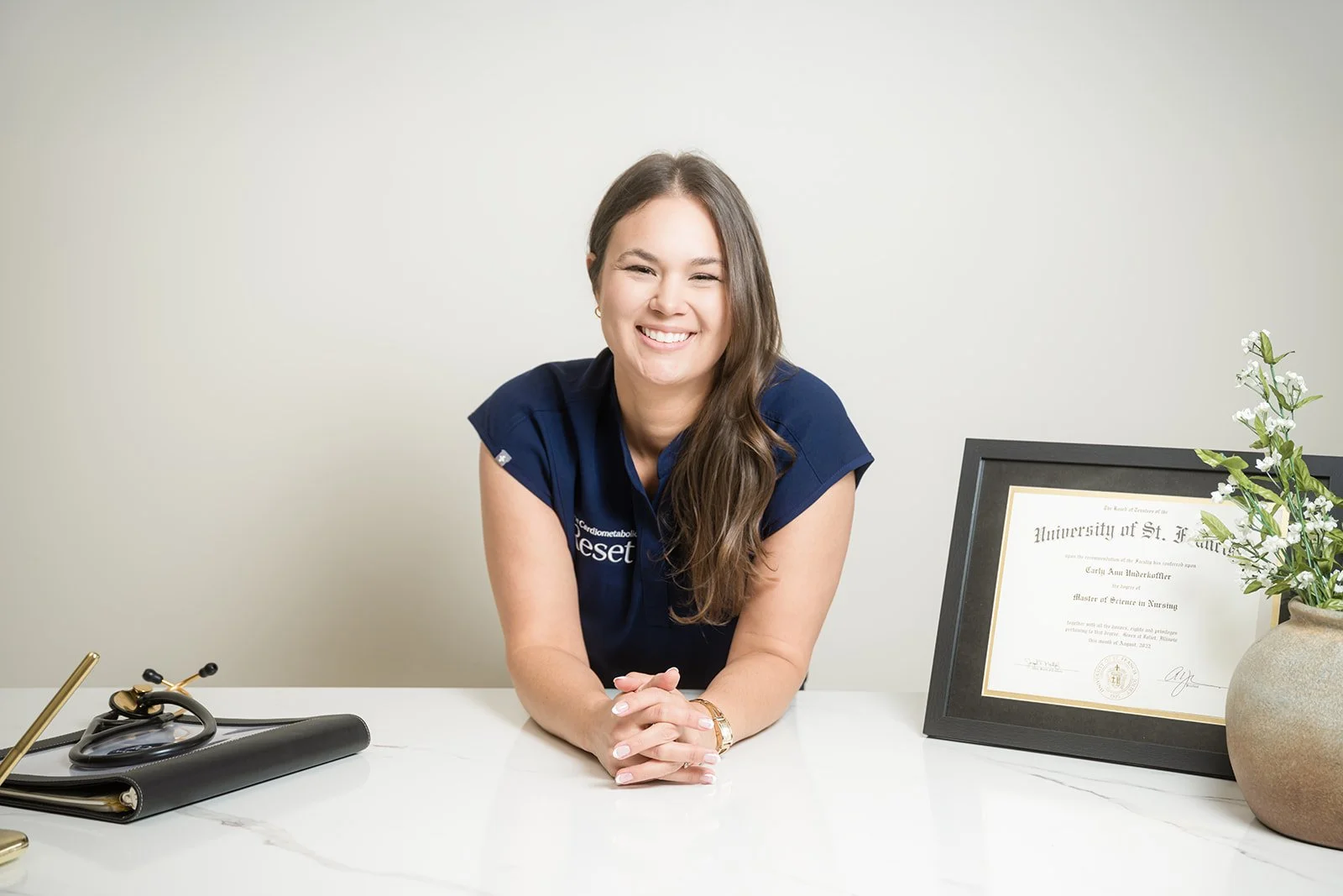 A woman with long brown hair smiling at a desk with a framed diploma, a potted plant, and a stethoscope.