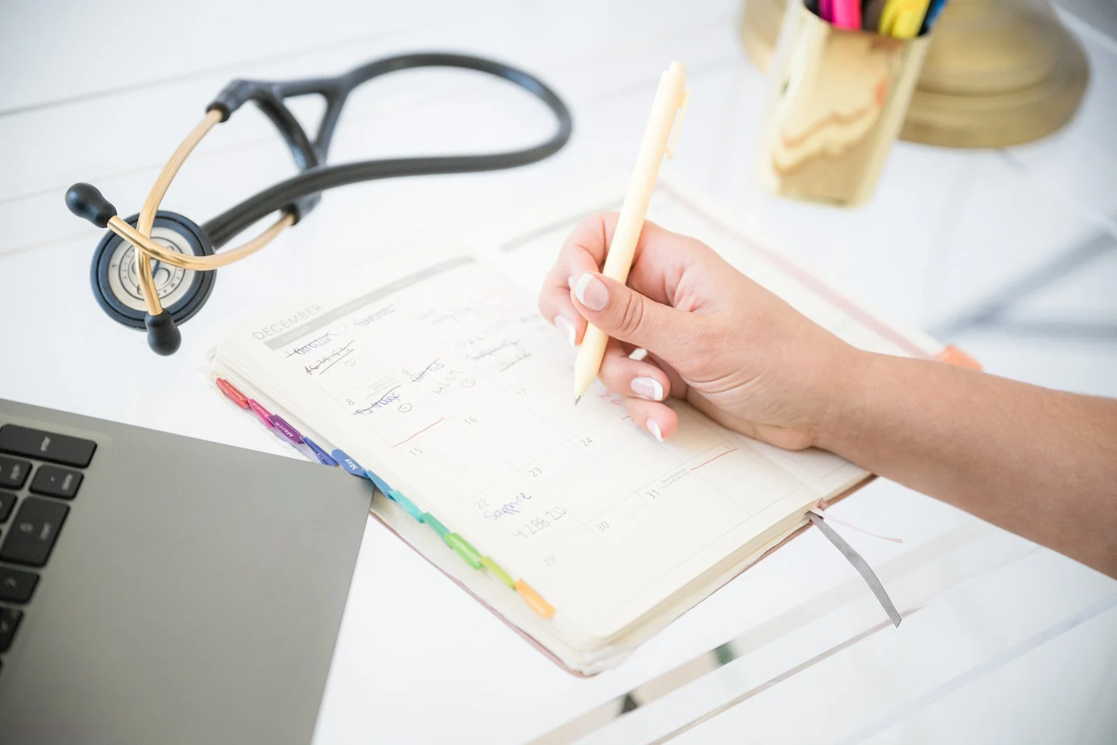 A person writing in a planner, surrounded by a laptop, a stethoscope, and a container of colorful pens on a white desk.