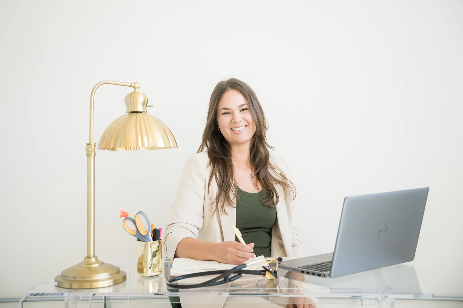 A woman sitting at a desk smiling, with a laptop, notebook, pen, stethoscope, a gold desk lamp, and a pen holder with scissors and markers, against a plain white wall.