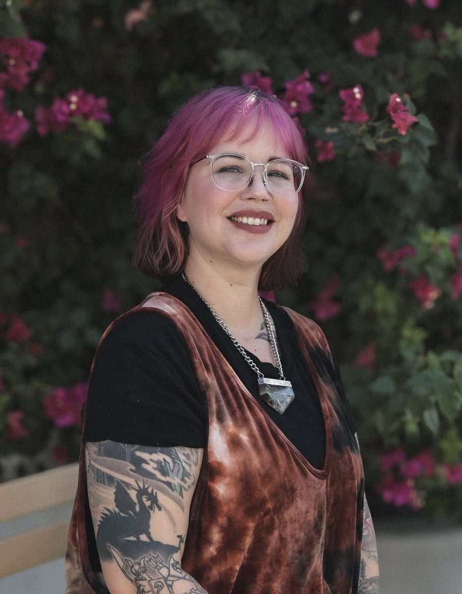 A young woman with pink and purple hair, wearing glasses, smiling, standing outdoors near pink flowering bushes, dressed in a black shirt, tie-dye brown and black shirt, with tattoos on her arms, wearing a large pendant necklace.