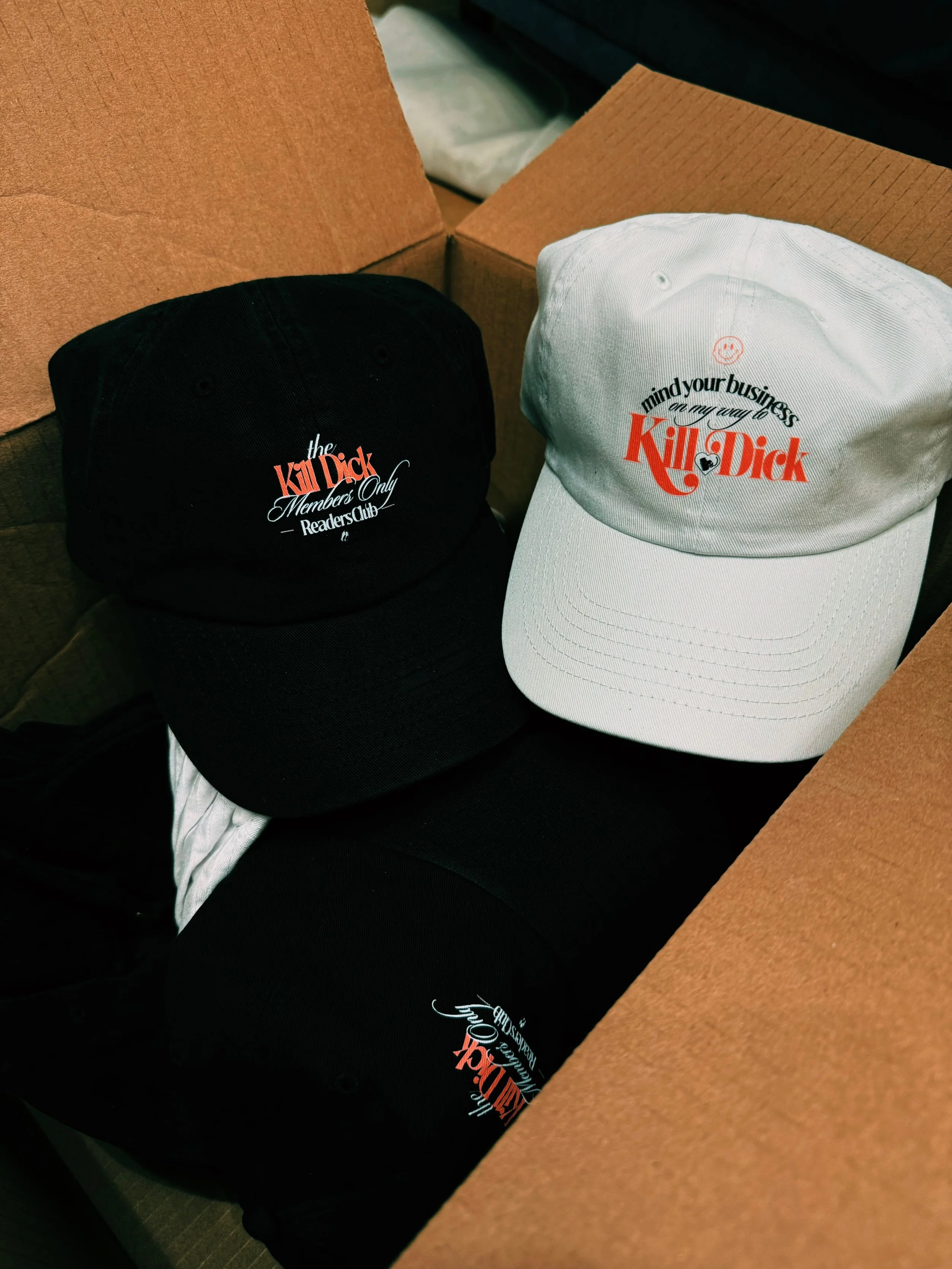 Three caps with the phrase 'Kill Dick' and a smaller text that says 'Members Only Readers Club' and 'mind your business on my way to Kill Dick'; two black caps and one white cap in a cardboard box.