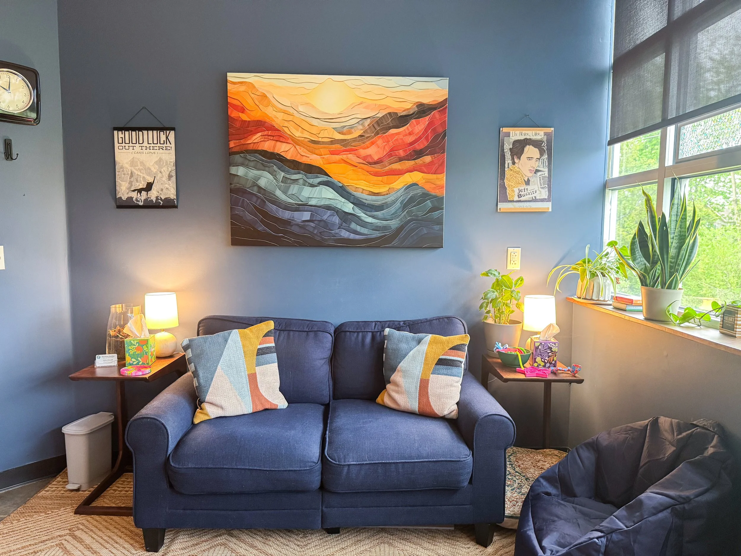A cozy living room corner with a blue couch, colorful geometric pillows, and a wooden side table with a plant, lamps, and decorative objects. The wall features a large abstract landscape painting with warm sunset colors, and smaller posters. Sunlight streams through window blinds, illuminating the space.