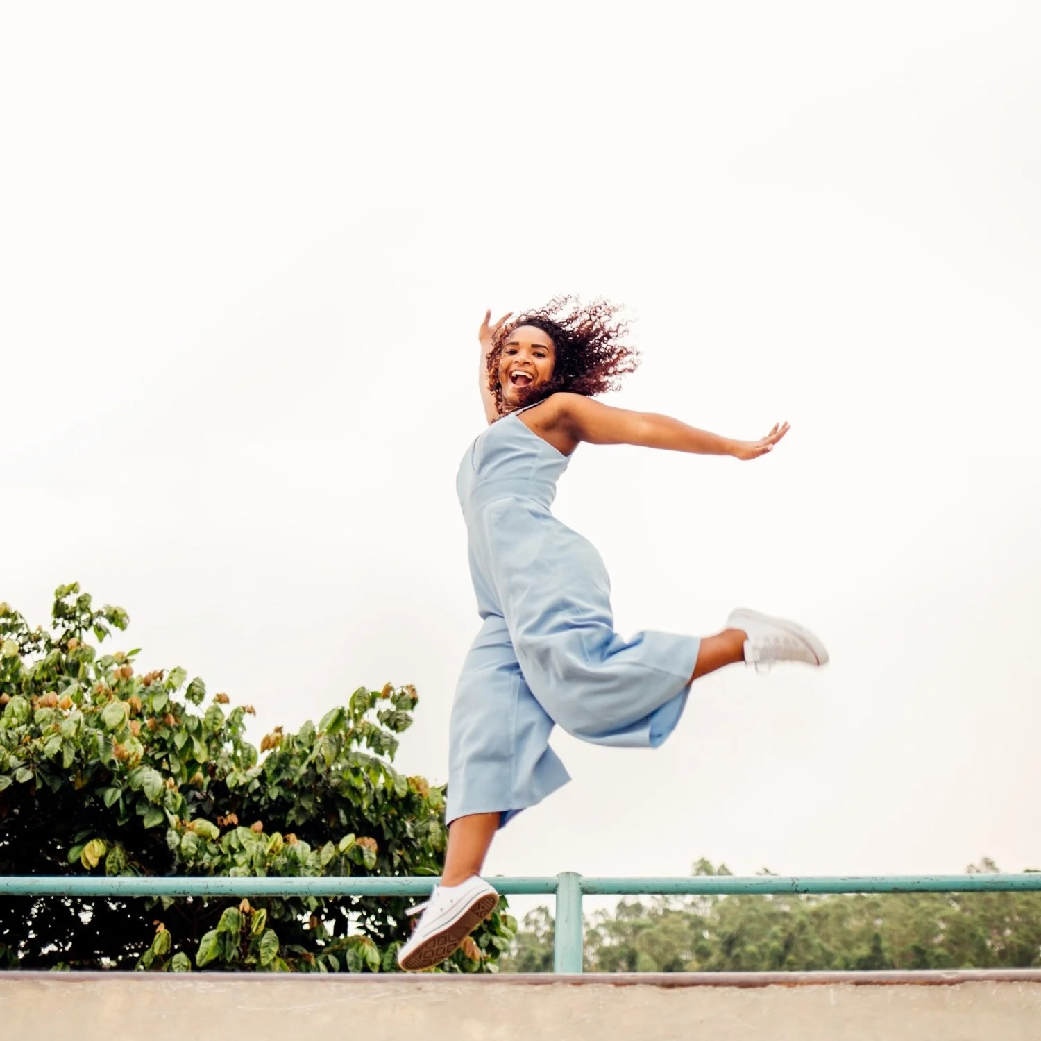 A woman in a light blue dress and white sneakers jumping in the air on a sunny day with a bush and trees in the background.