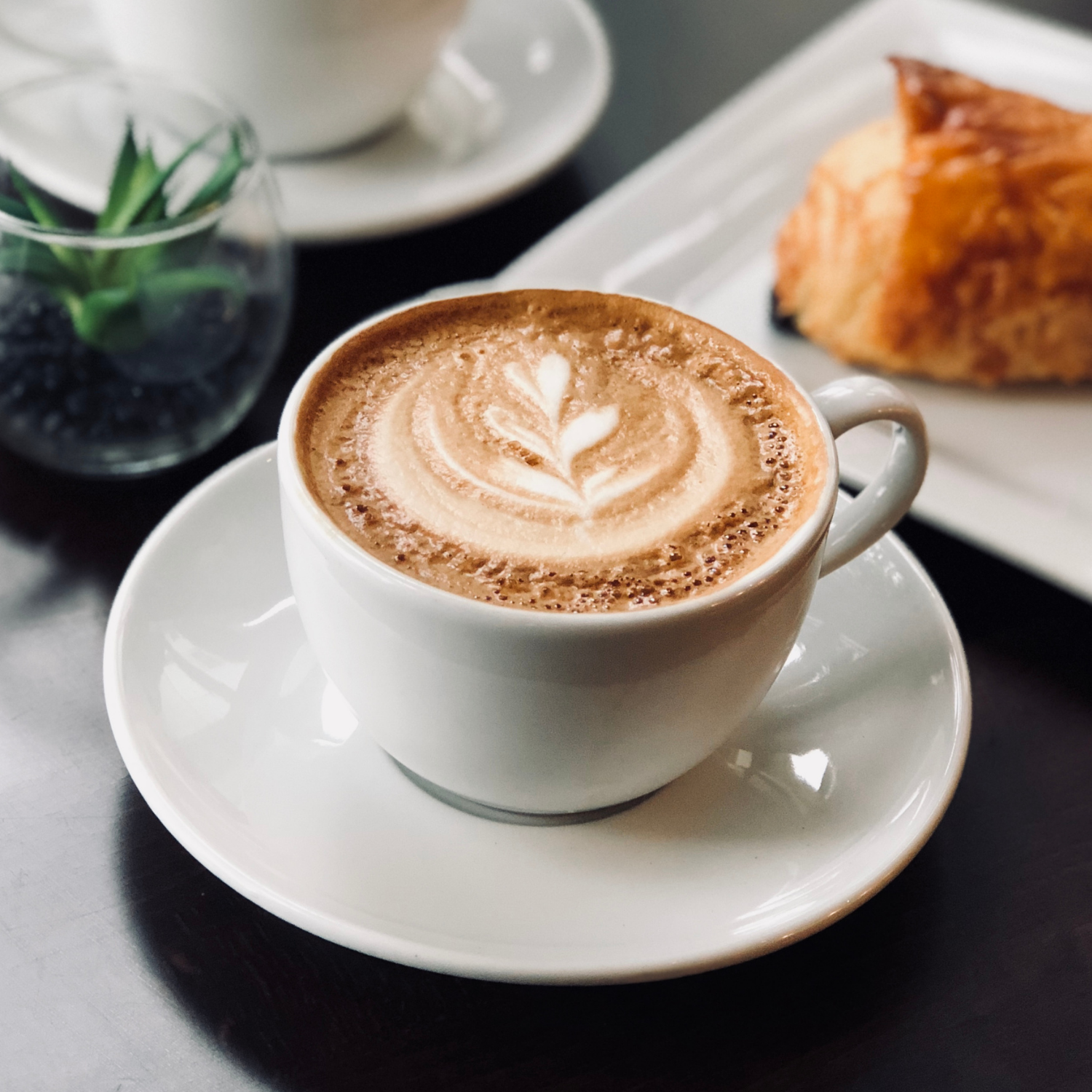 A cup of coffee with latte art on top, served on a white saucer, with a pastry on a white plate in the background.
