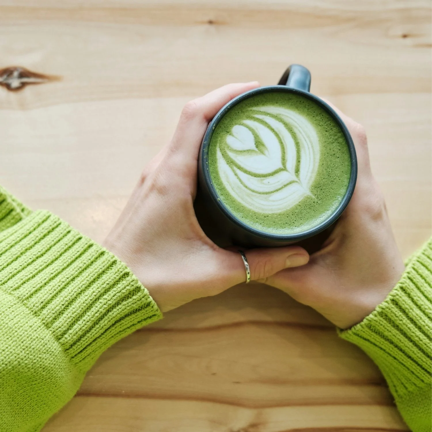 Person holding a black mug with green matcha latte with latte art on a wooden table, wearing a neon green sweater.