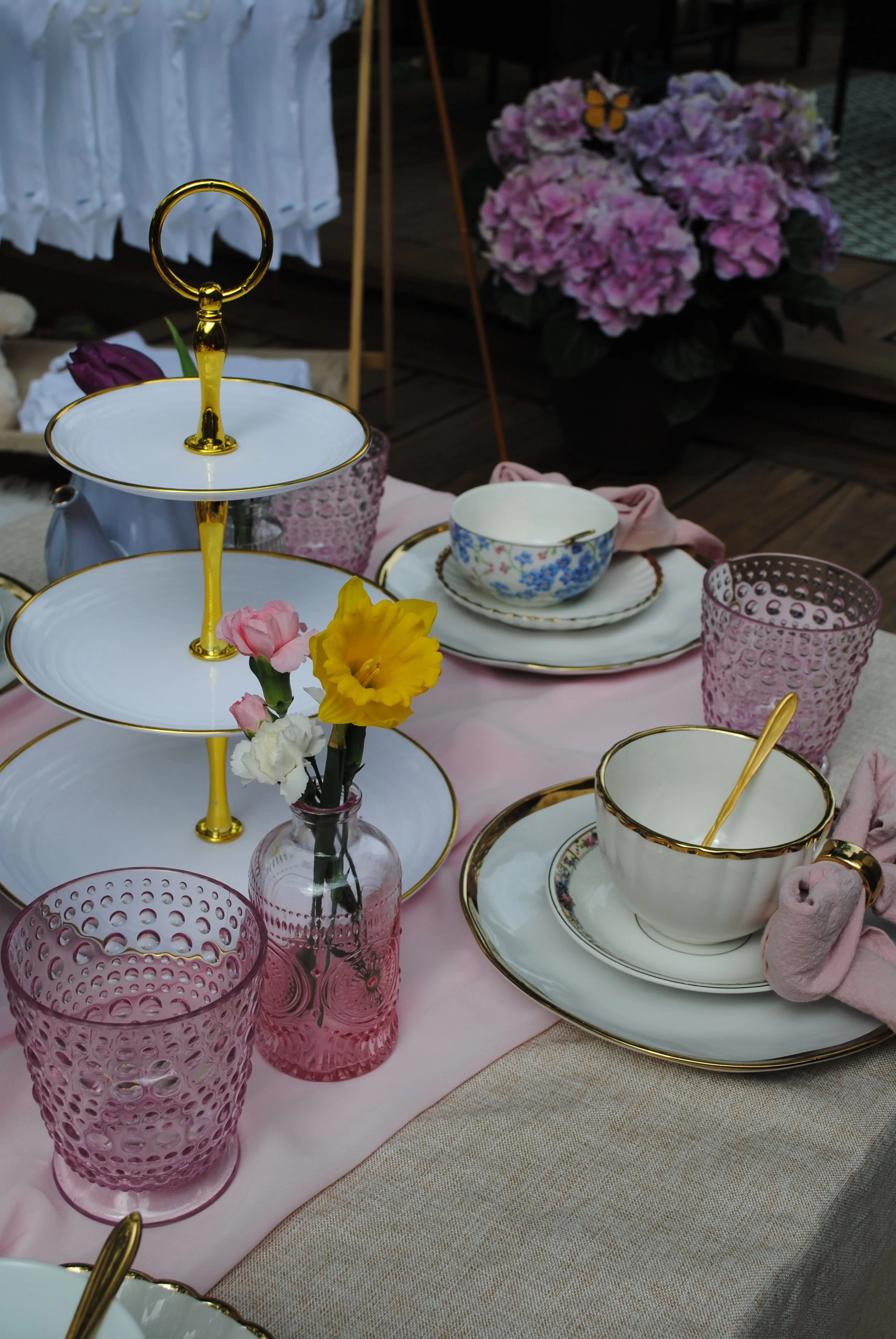 A table set for tea with pink tablecloth, floral china cups and saucers, pink and purple textured glasses, a small pink vase with yellow, pink, and white flowers, and a tiered serving stand with white plates and golden accents. In the background, there are pink hydrangea flowers and a butterfly decoration.