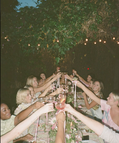 People sitting at a long outdoor dinner table, raising glasses in a toast under string lights at night.