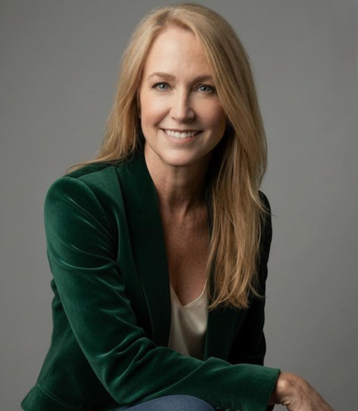 Portrait of a woman with long blonde hair, wearing a green blazer and a white top, smiling at the camera against a gray background.