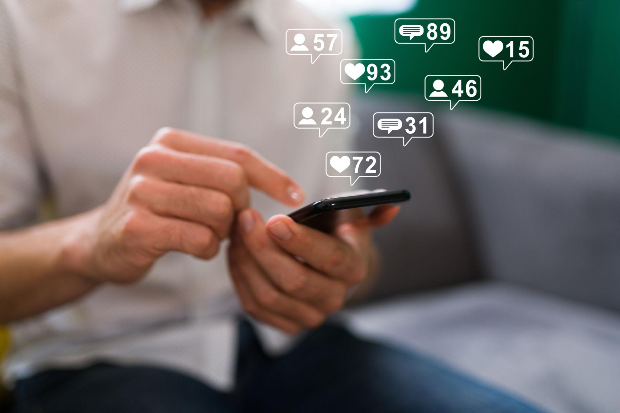 Person holding a smartphone with social media notification icons floating above it, showing likes, comments, and followers.