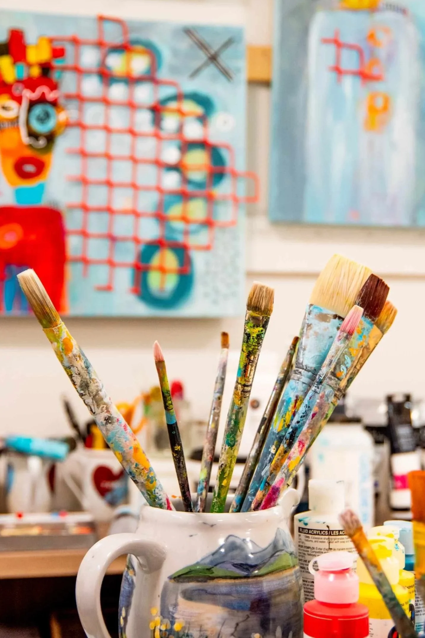 Paintbrushes in a white ceramic mug, with a colorful painter's studio in the background.