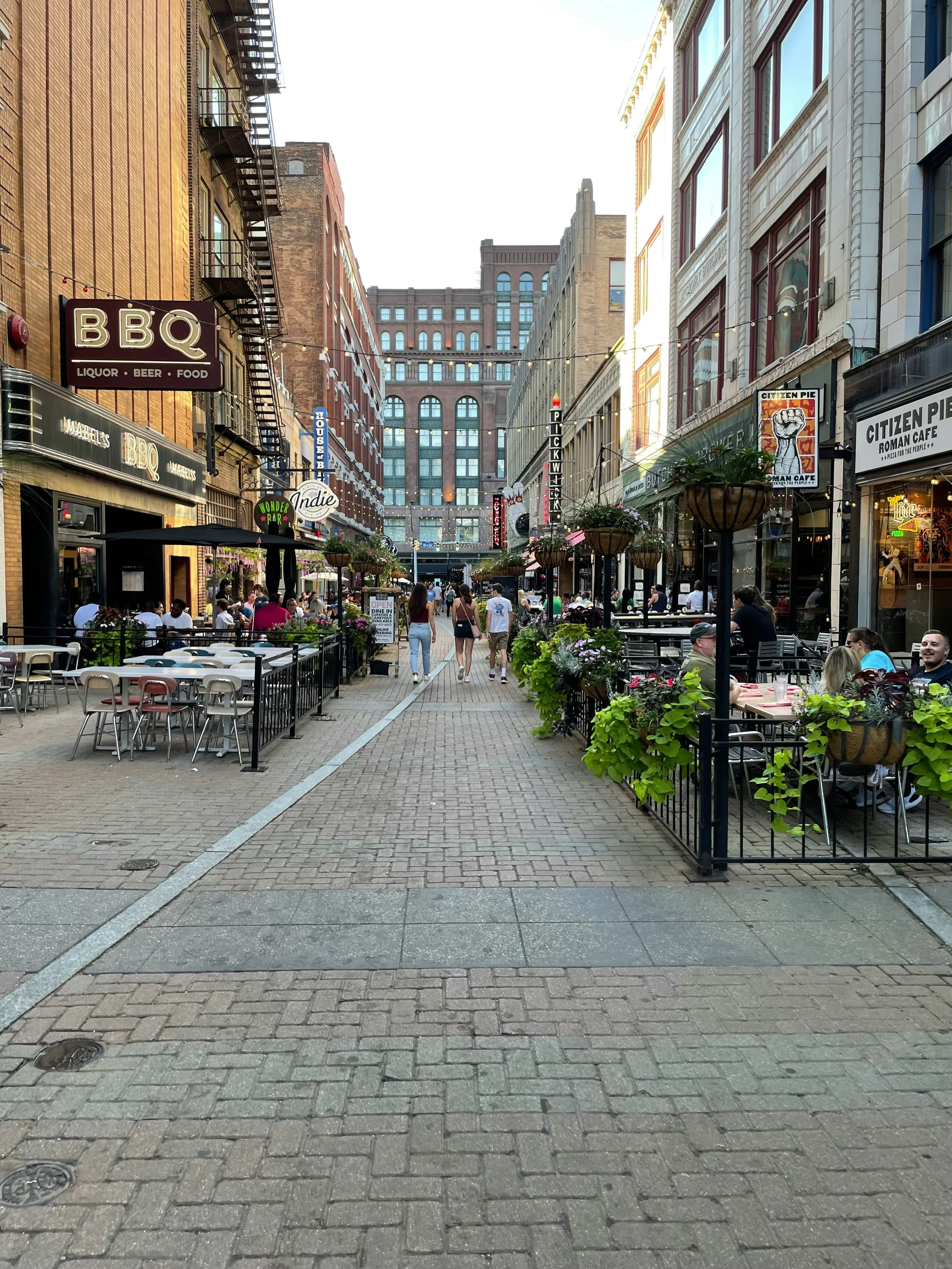Outdoor pedestrian street with cafes and restaurants, people walking and sitting, decorated with hanging flower baskets, tall buildings in the background.