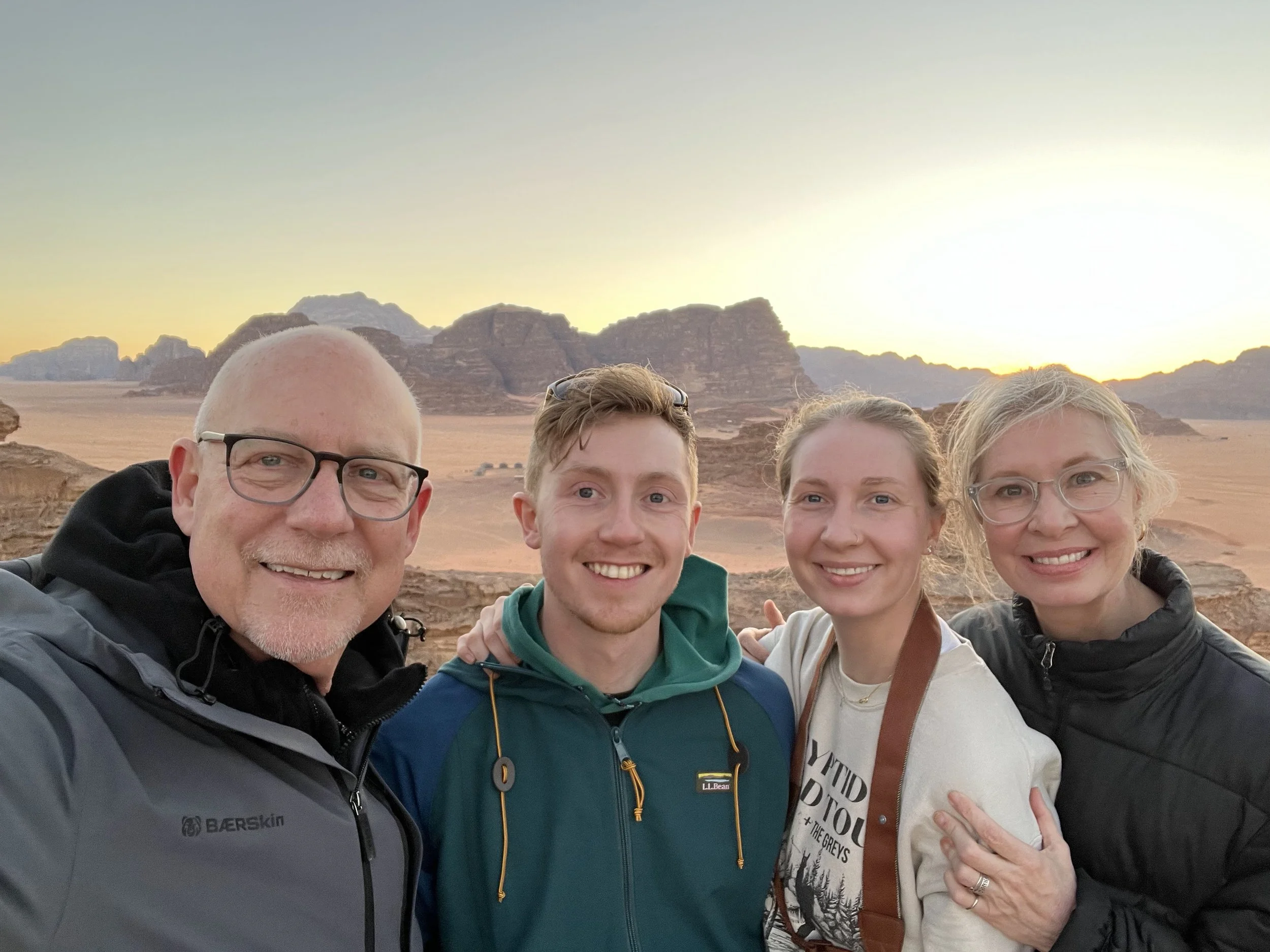 Four people smiling and taking a selfie together in a desert landscape at sunset, with rocky formations in the background.