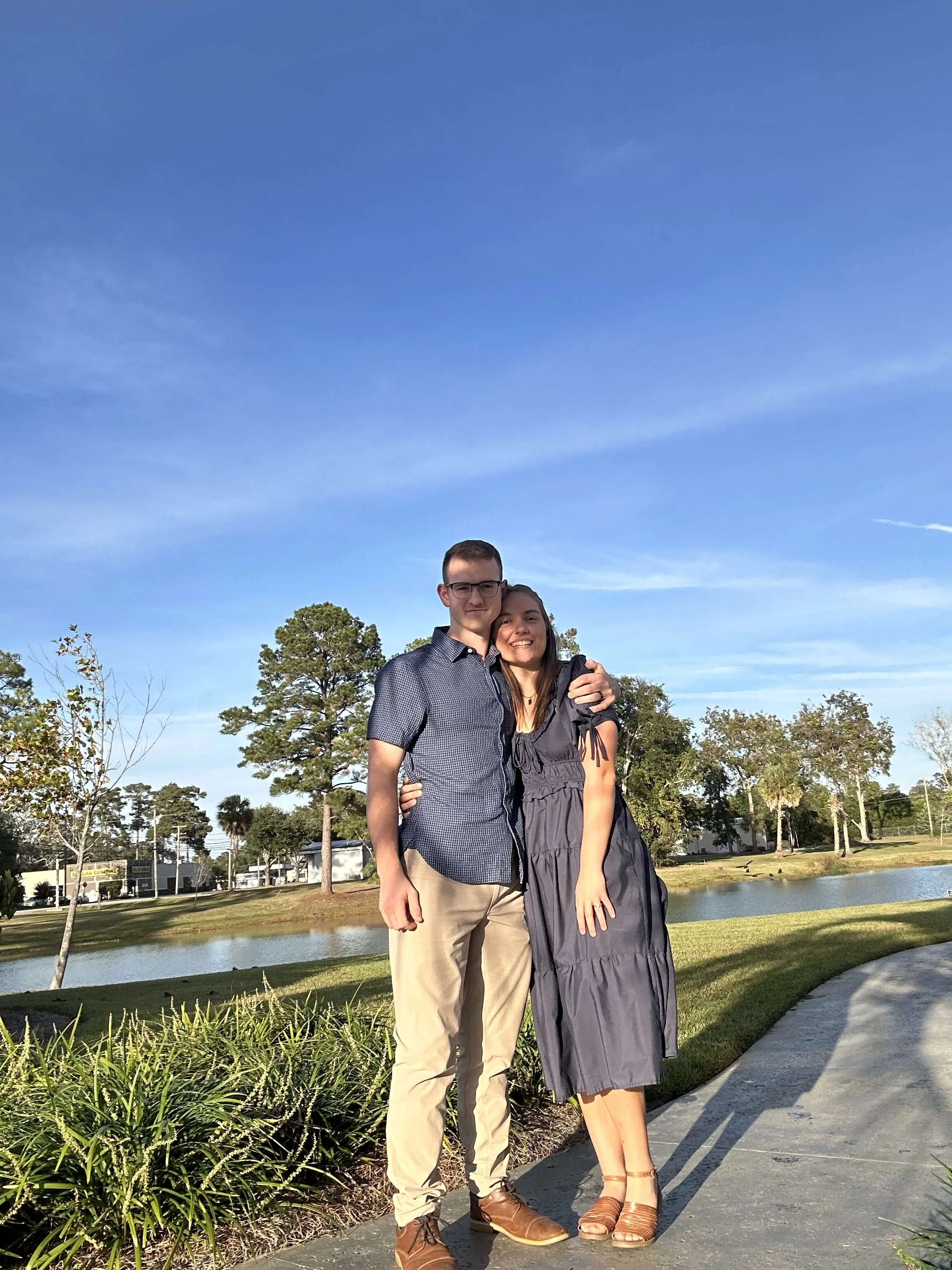 A young couple standing outdoors near a pond, smiling and hugging, with trees and a blue sky in the background.