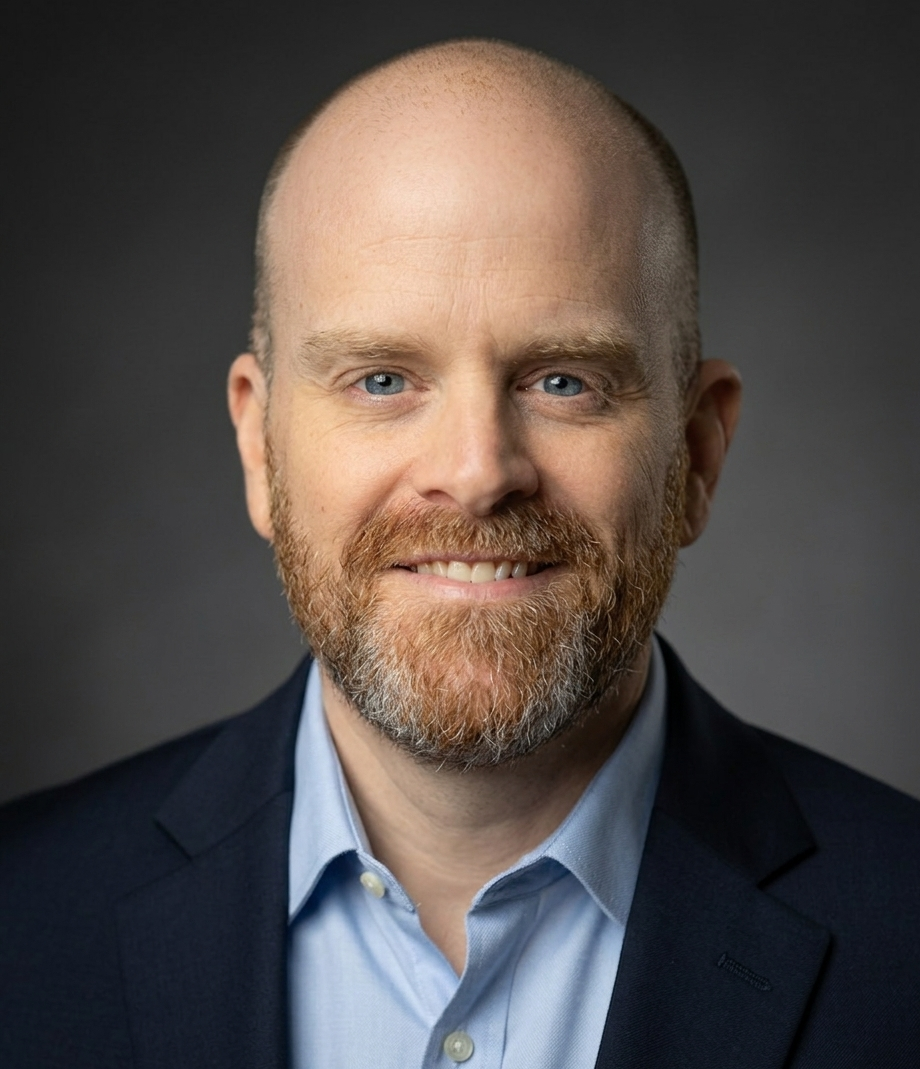 A portrait of a smiling man with a beard and blue eyes, wearing a dark blazer and light blue collared shirt against a dark background.