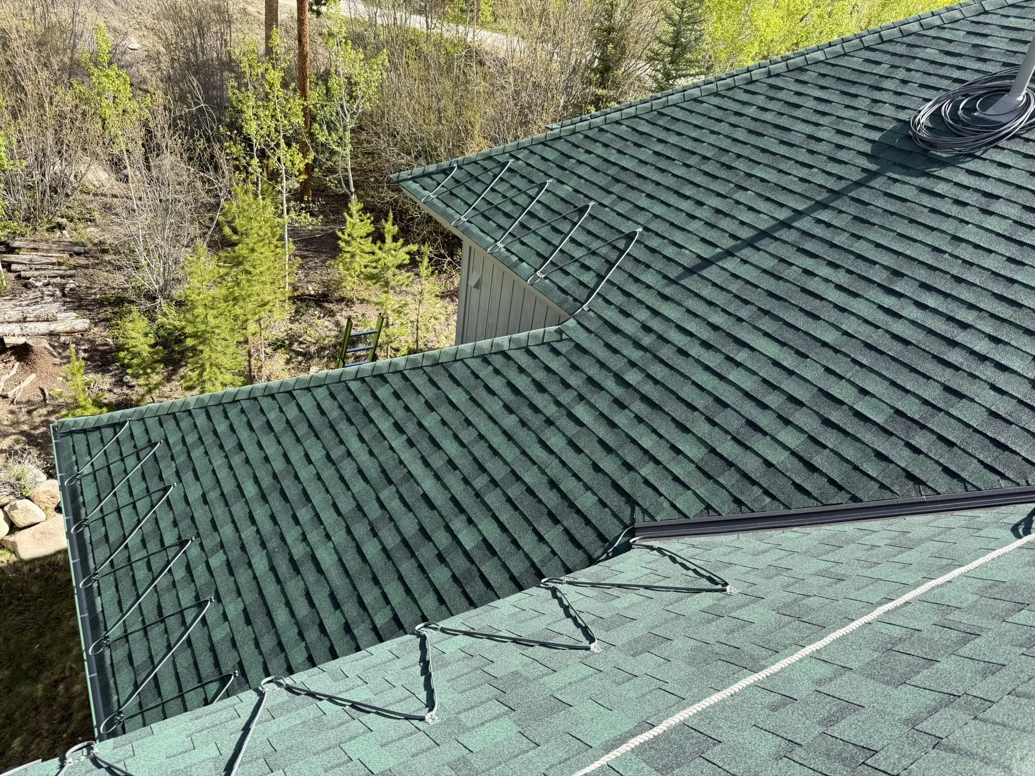 Heat trace cable along the eaves and valleys of a green shingle roof surrounded by pine trees