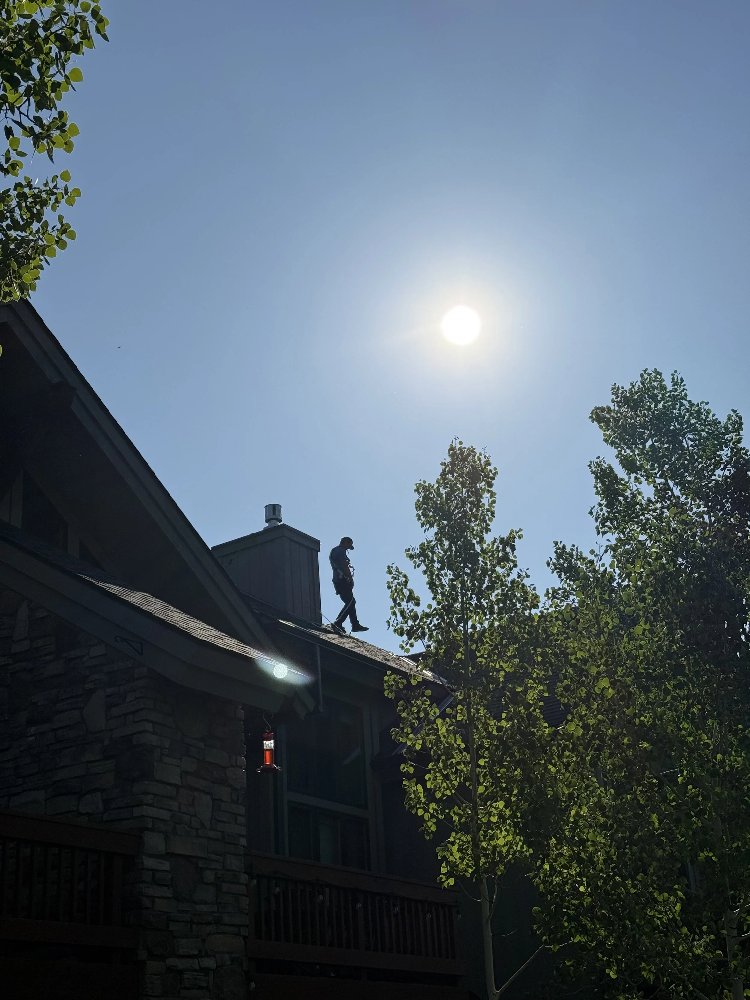 A person walking on the roof of a house under a bright sun in a clear blue sky, with trees surrounding the house.