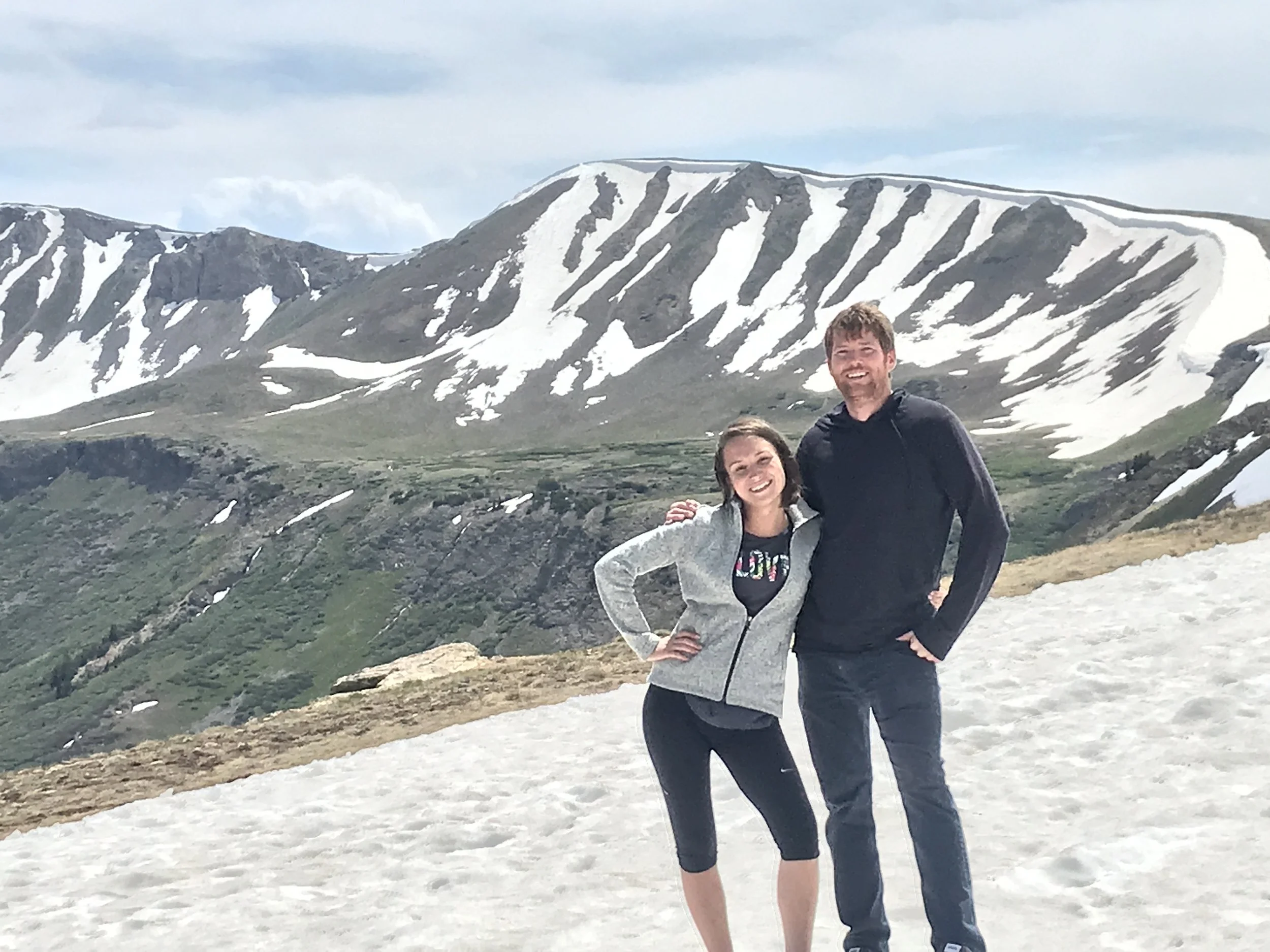 A smiling couple stands on a snow-covered mountainside with snow-capped peaks in the background.