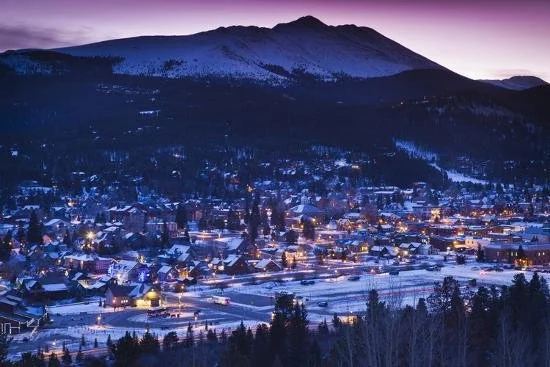 A winter mountain town at dusk with snow-covered buildings and streets, and a mountain in the background.