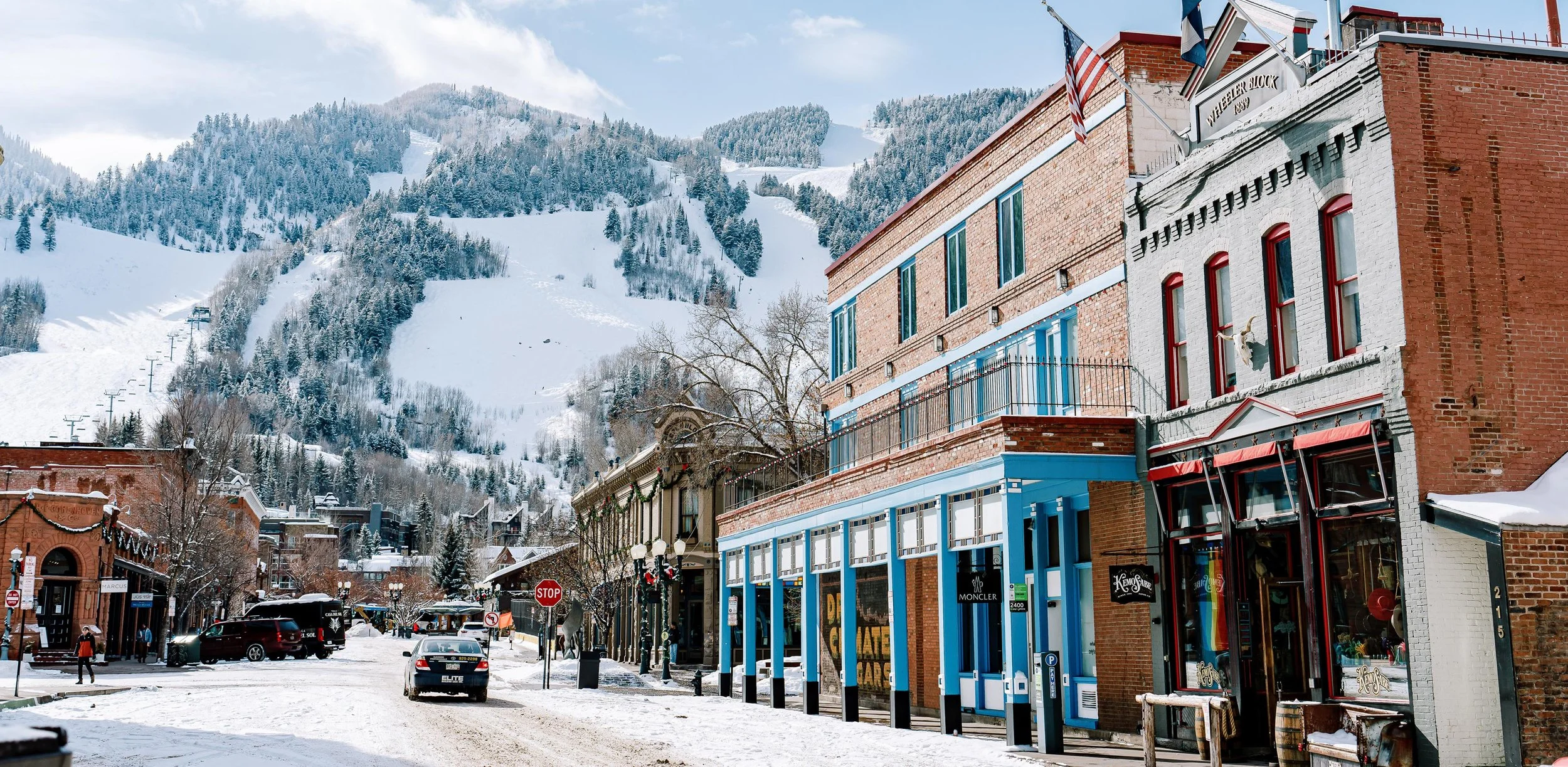 Snow-covered mountain town street with colorful storefronts, leafless trees, parked cars, and ski slopes on the mountain in the background.