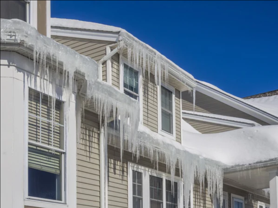 A house with icicles hanging from the roof and snow on the roof under a clear blue sky.
