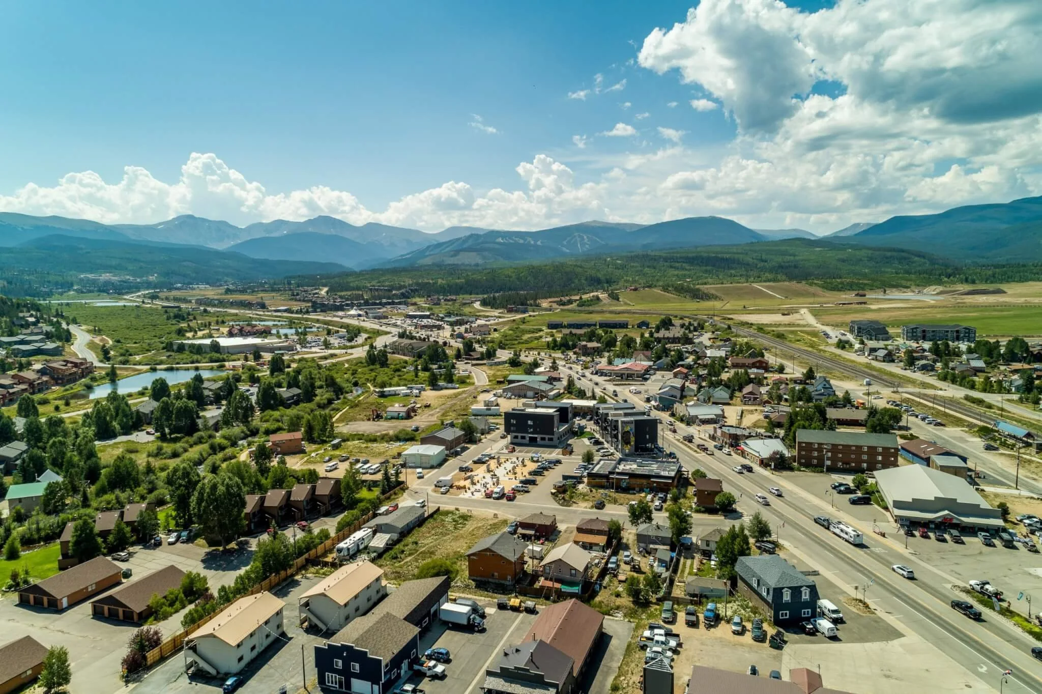 Aerial view of a small town surrounded by mountains, with buildings, roads, and a body of water visible in a lush green landscape under a partly cloudy sky.