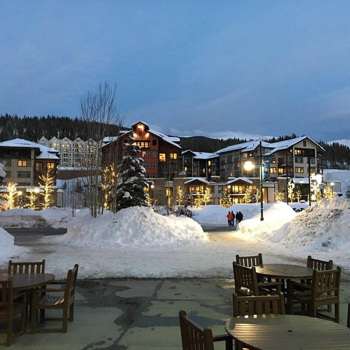 Snow-covered outdoor patio with wooden tables and chairs, snow piled along the walkway, buildings with lit windows and string lights, and a winter landscape with trees in the background.